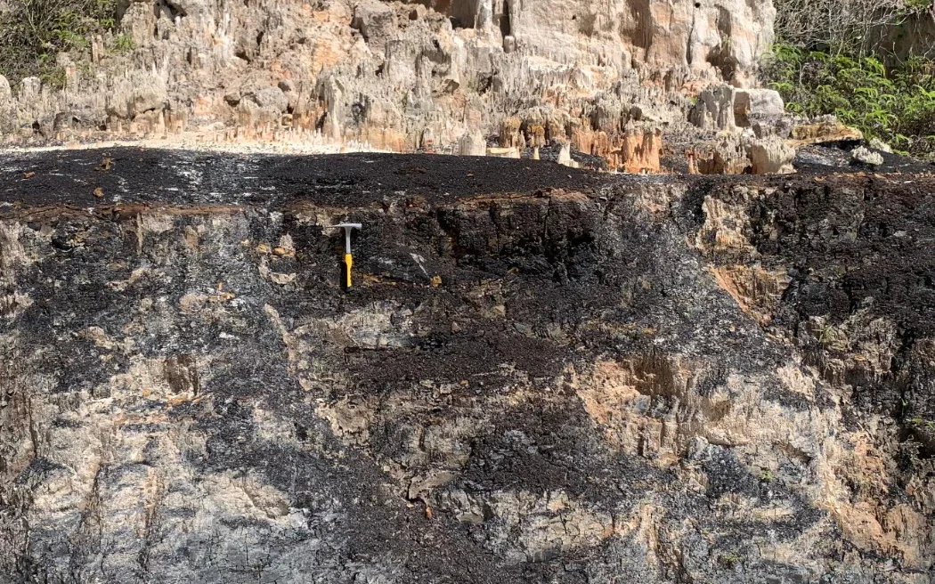 A rock hammer for scale against a dark rock face below a pale cliff