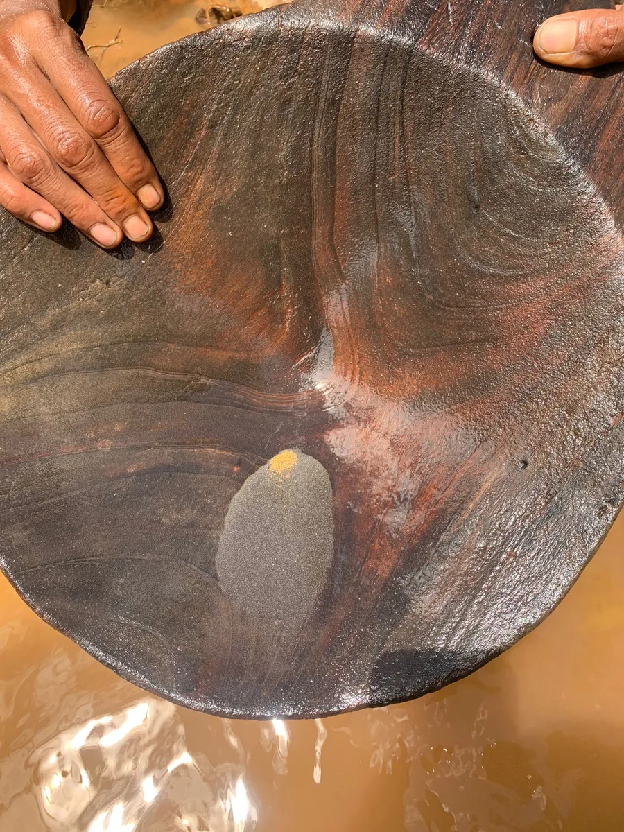 Hands holding a wooden pan with fine dark sampled material at a stream