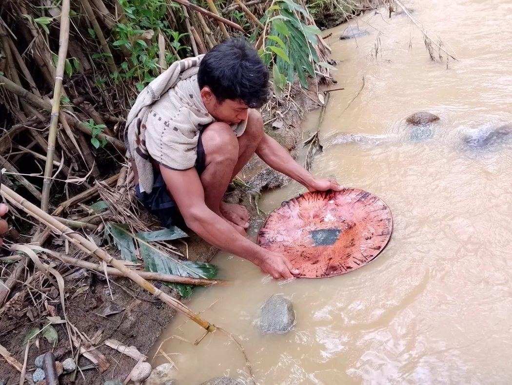 A person crouching at the edge of a muddy stream holding a wooden pan